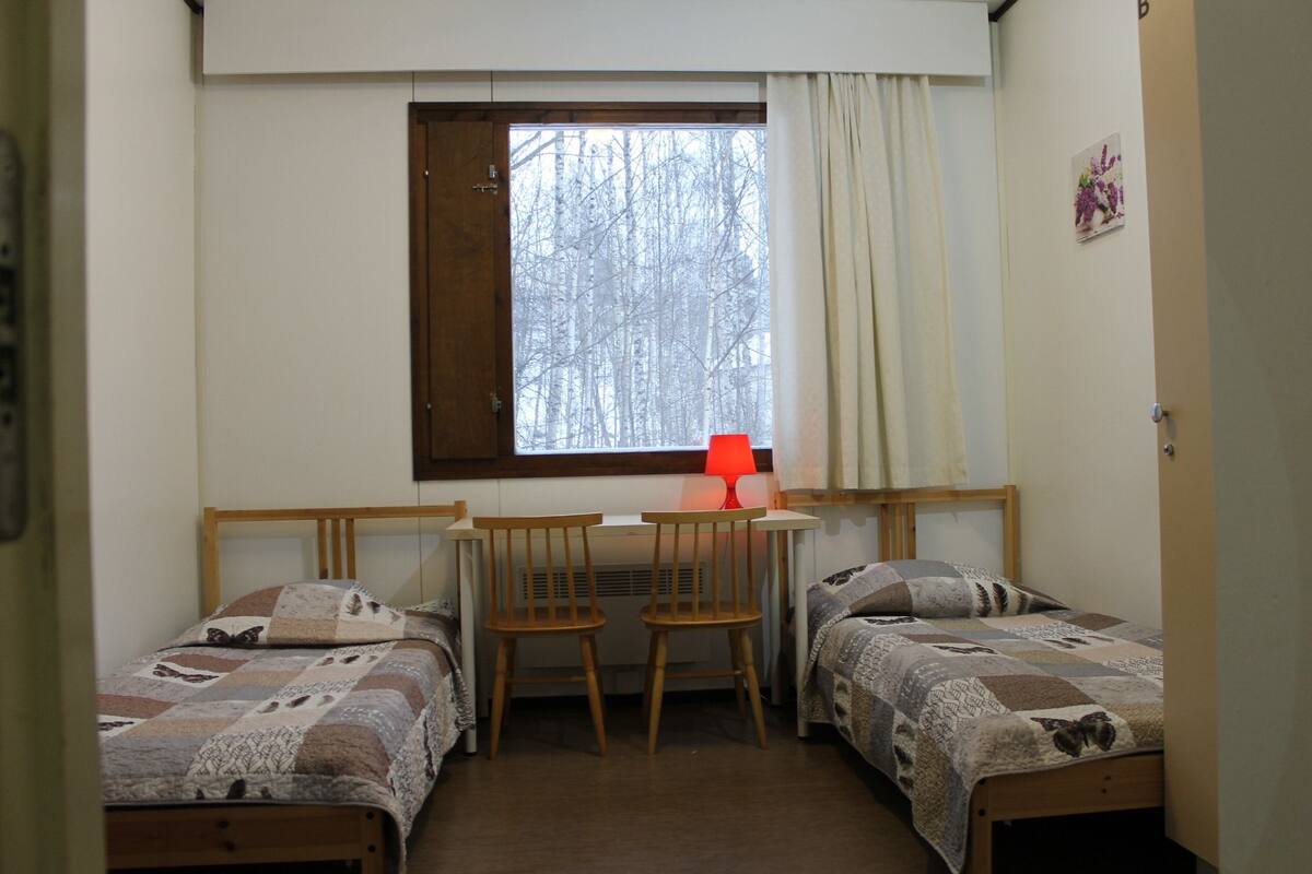 Une petite chambre confortable avec deux lits simples, un bureau en bois et une chaise. Il y a une fenêtre avec un rideau et une lampe rouge sur le bureau.