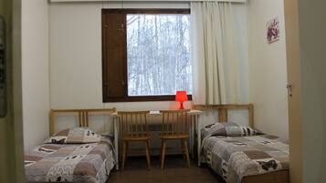 A small, cozy room with two single beds, a wooden desk, and a chair. There is a window with a curtain, and a red lamp on the desk.