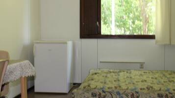A room with a bed, a small white refrigerator, a chair, and a window with a view of trees.