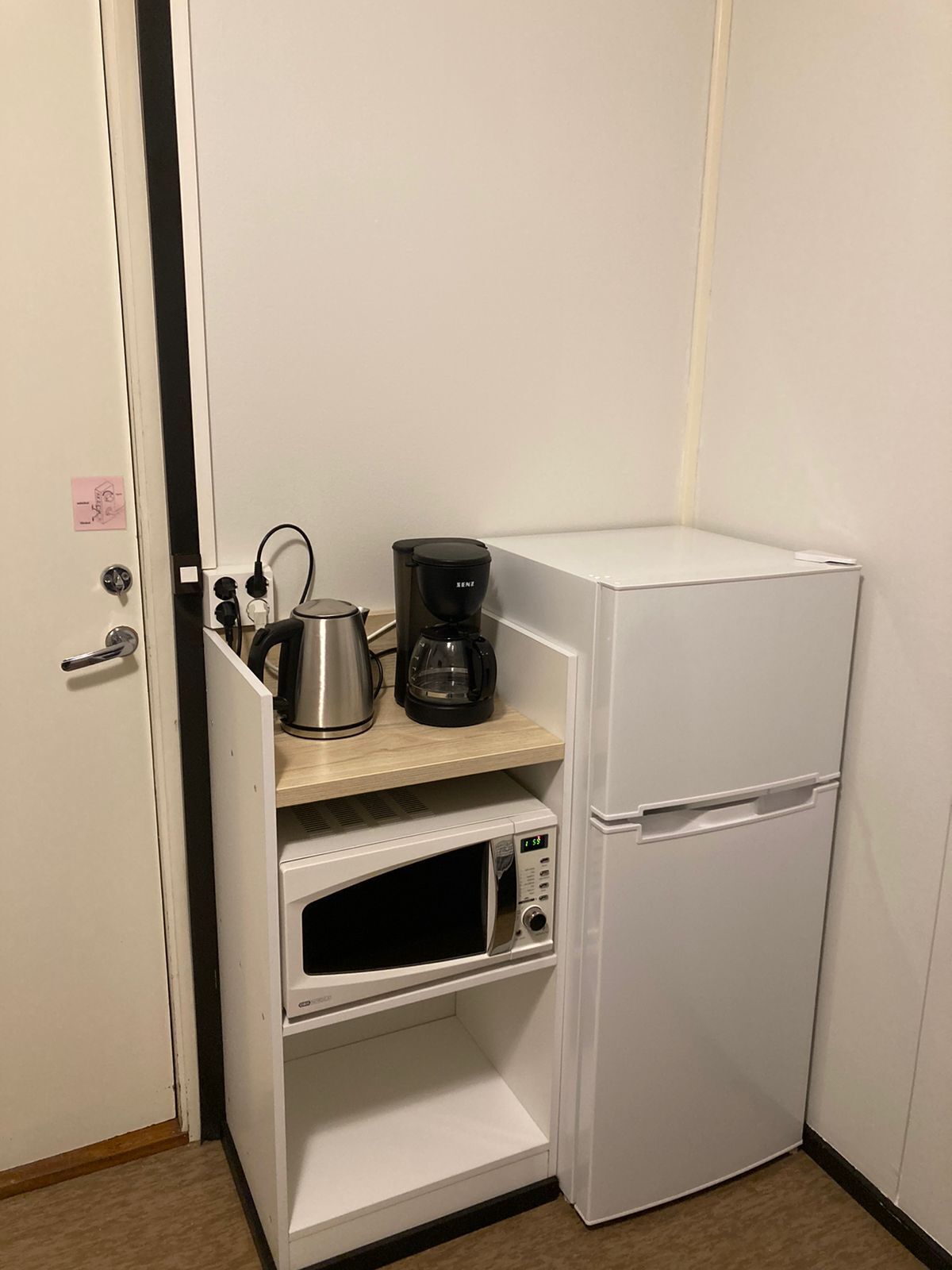 Una kitchenette compacta con microondas, cafetera y hervidor sobre una estantería, y un refrigerador.
