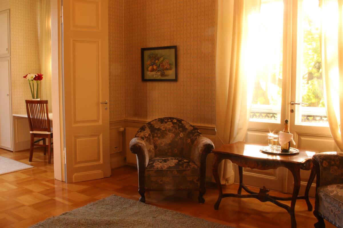 A room with wooden flooring, a floral armchair, a small table with a bottle and glasses, a chair, a vase with flowers, and a framed picture on the wall.
