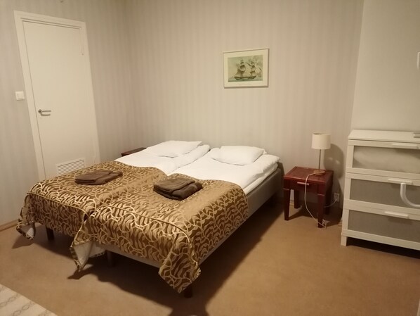 Una cama bien tendida con una colcha estampada y dos toallas, una mesita de noche con una lámpara, y un cuadro en la pared.