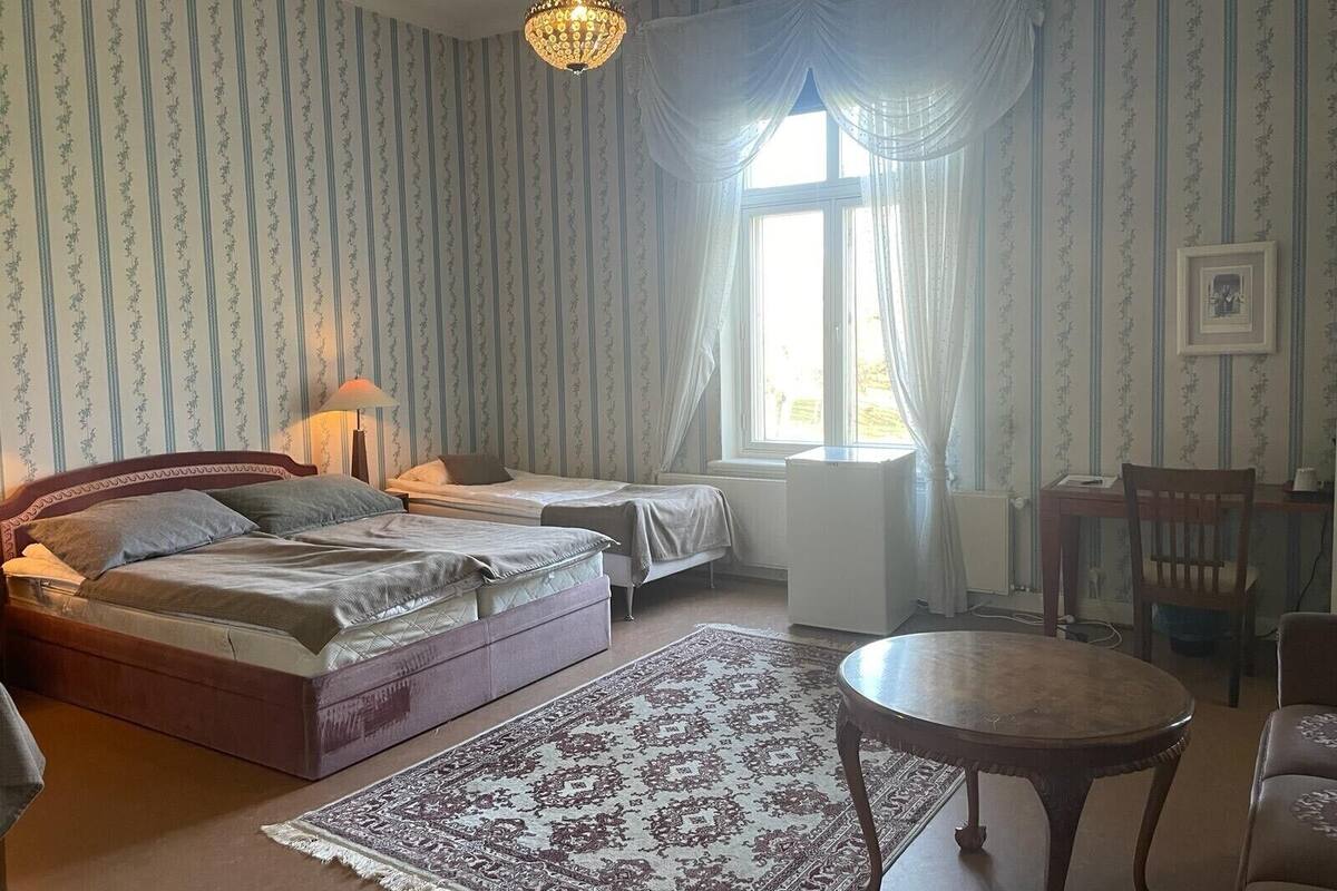 A room with two beds, a desk, a chair, a table, a chandelier, and a window with curtains.