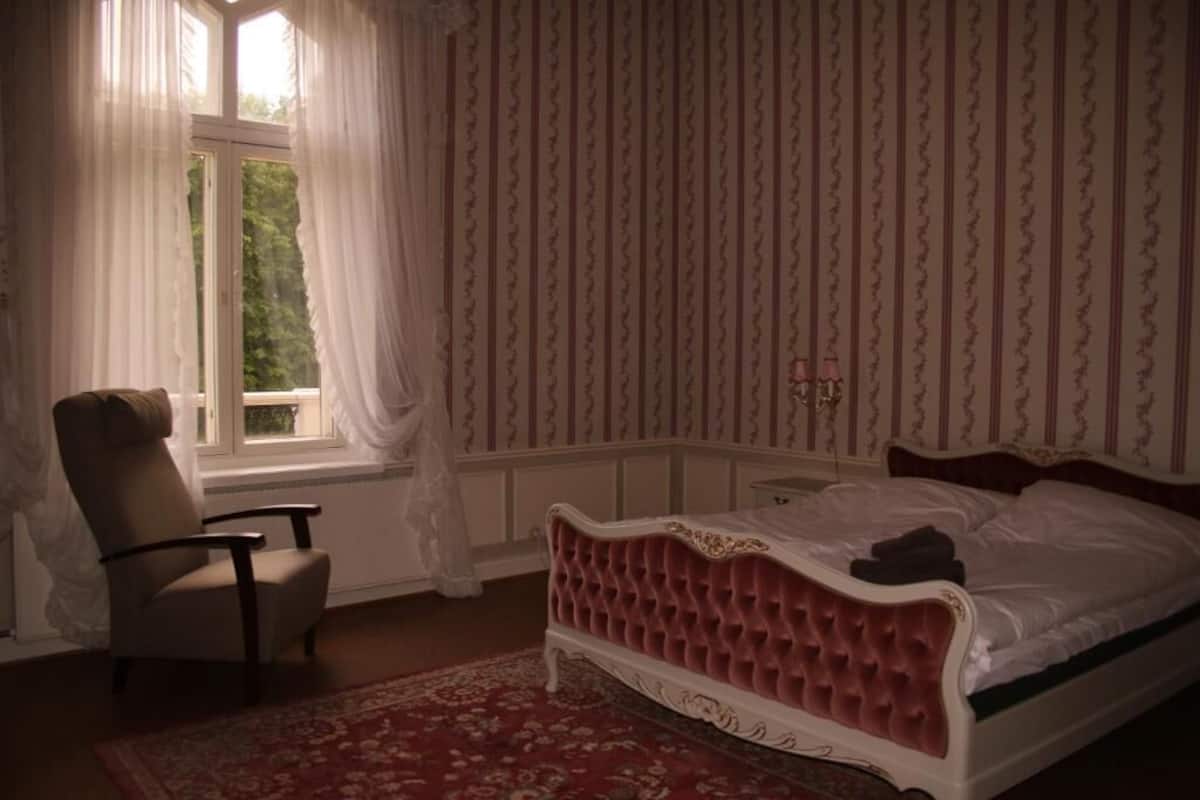 A bedroom with a large bed, a chair, a window with curtains, and a patterned wallpaper.