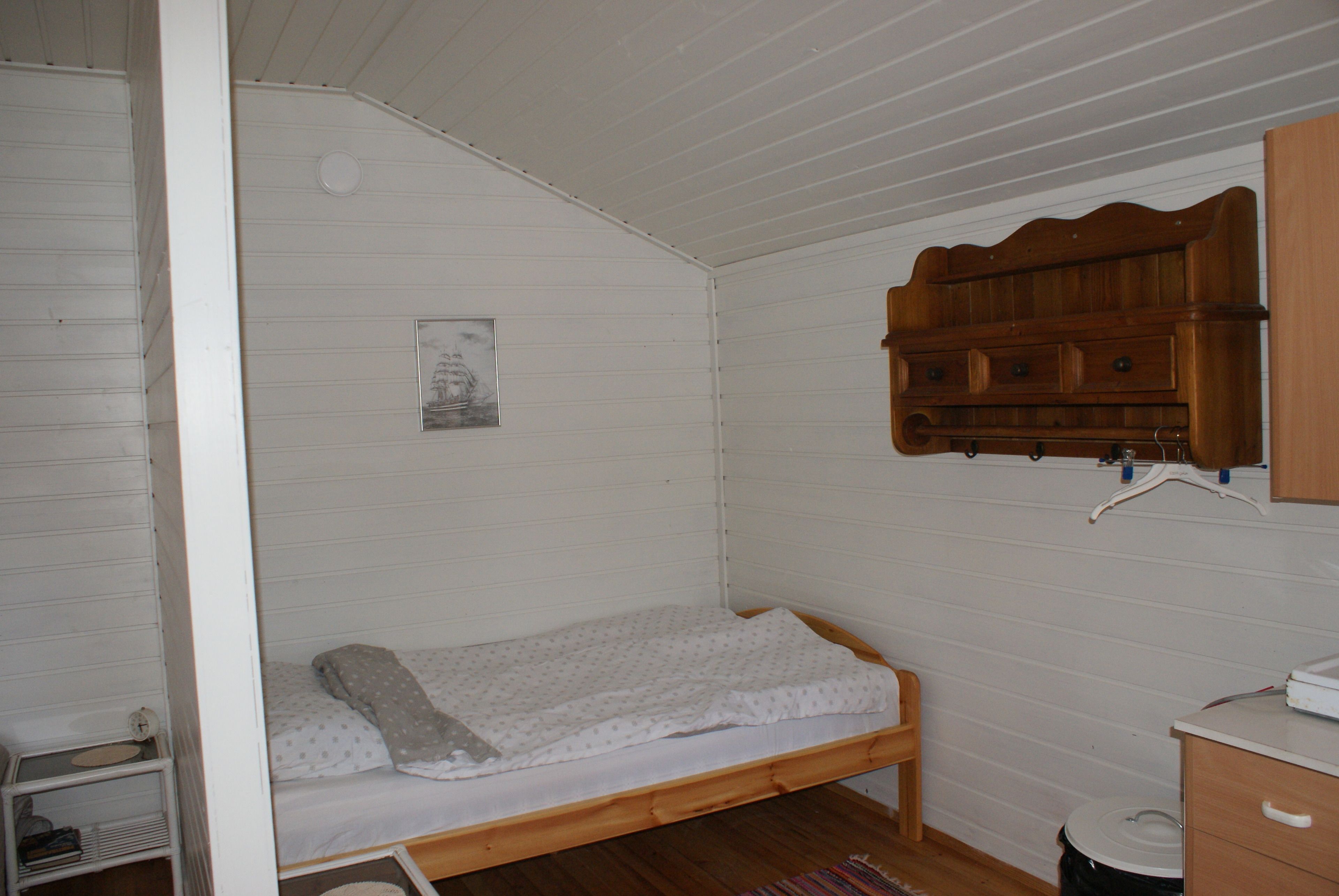 Una habitación pequeña con una cama individual tipo litera de madera, una pared blanca con un cuadro enmarcado, un armario de madera con objetos colgados y un estante pequeño.