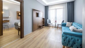 A modern living room with a blue sofa, a TV mounted on a wooden panel, a dining area with a table and chairs, and a large window with curtains. - Beyzas Hotel & Suites (Istanbul)