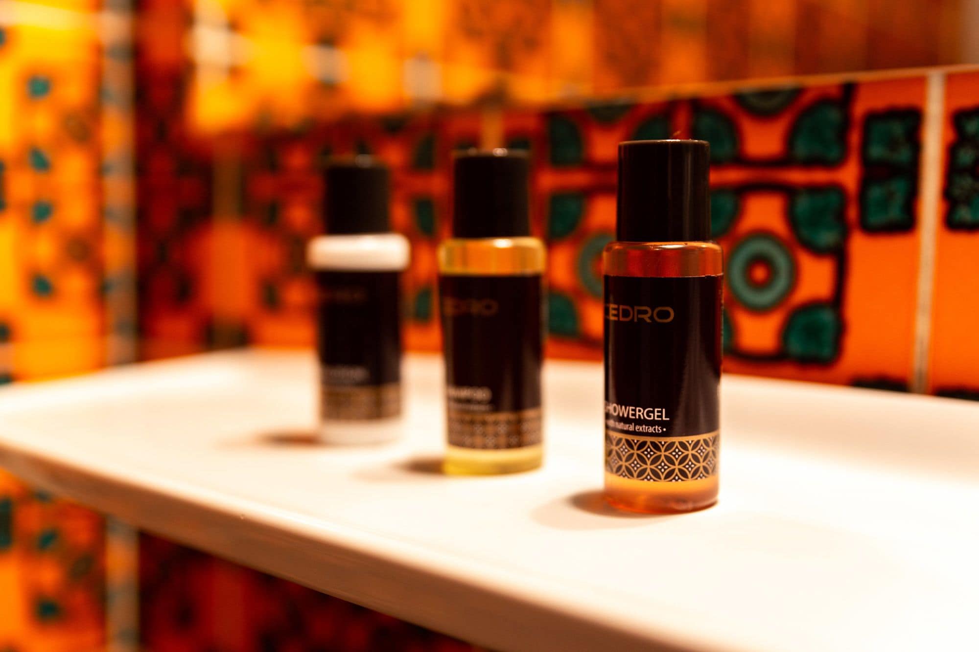 Three bottles of shower gel on a white surface with a colorful tiled wall in the background.