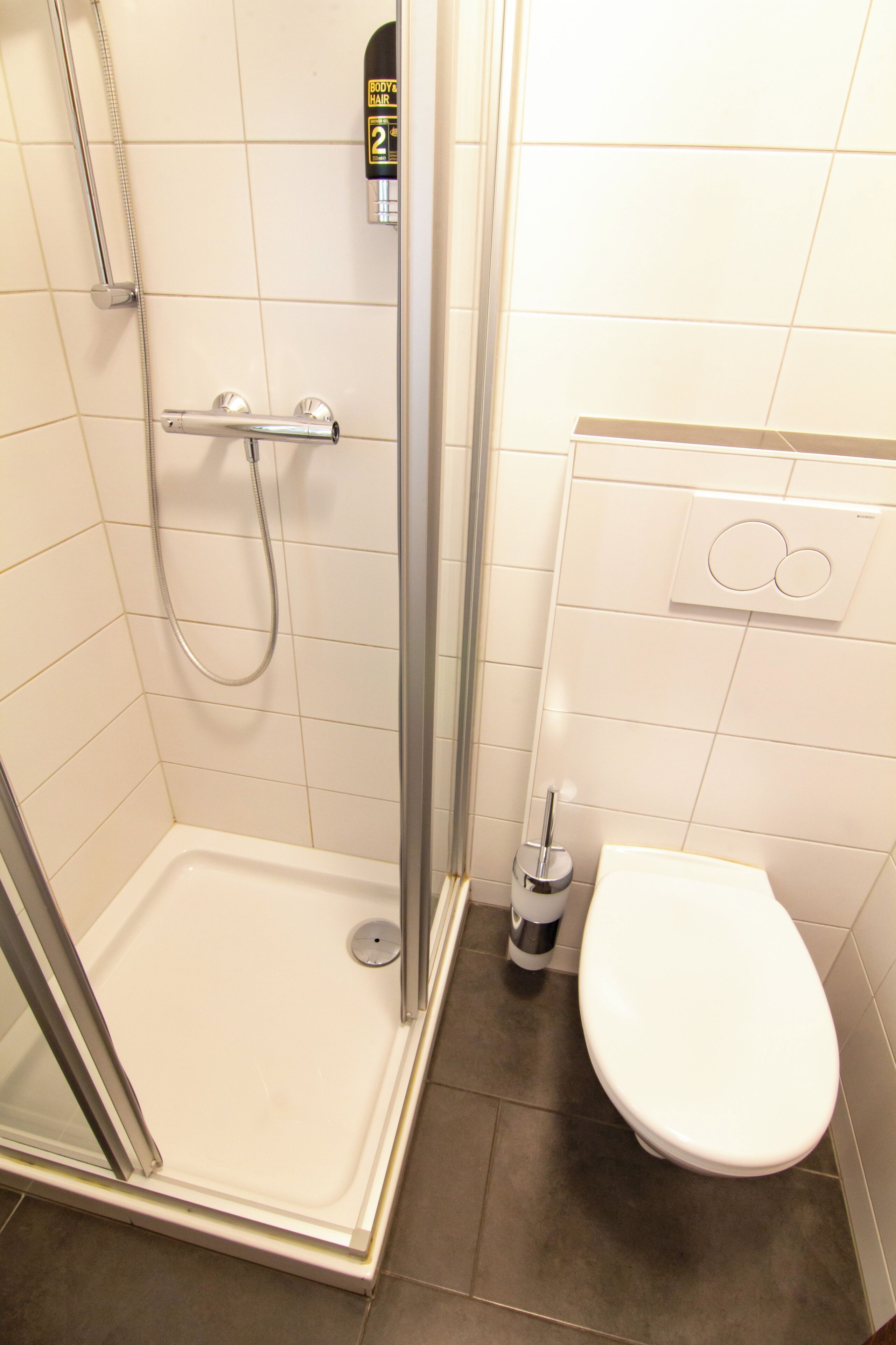 A shower with a glass door, a toilet, and a soap dispenser on the wall.