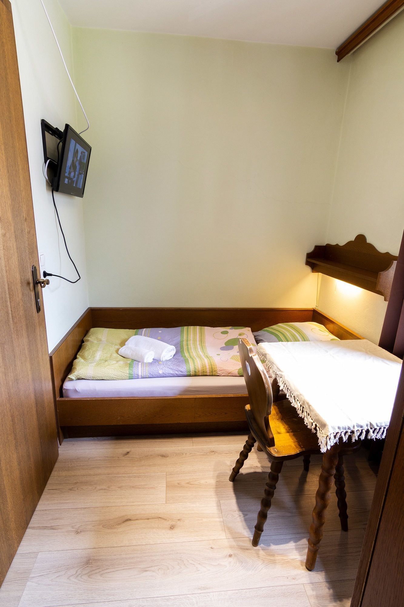 A small, compact room with a bed, a desk, and a chair. There is a television mounted on the wall.