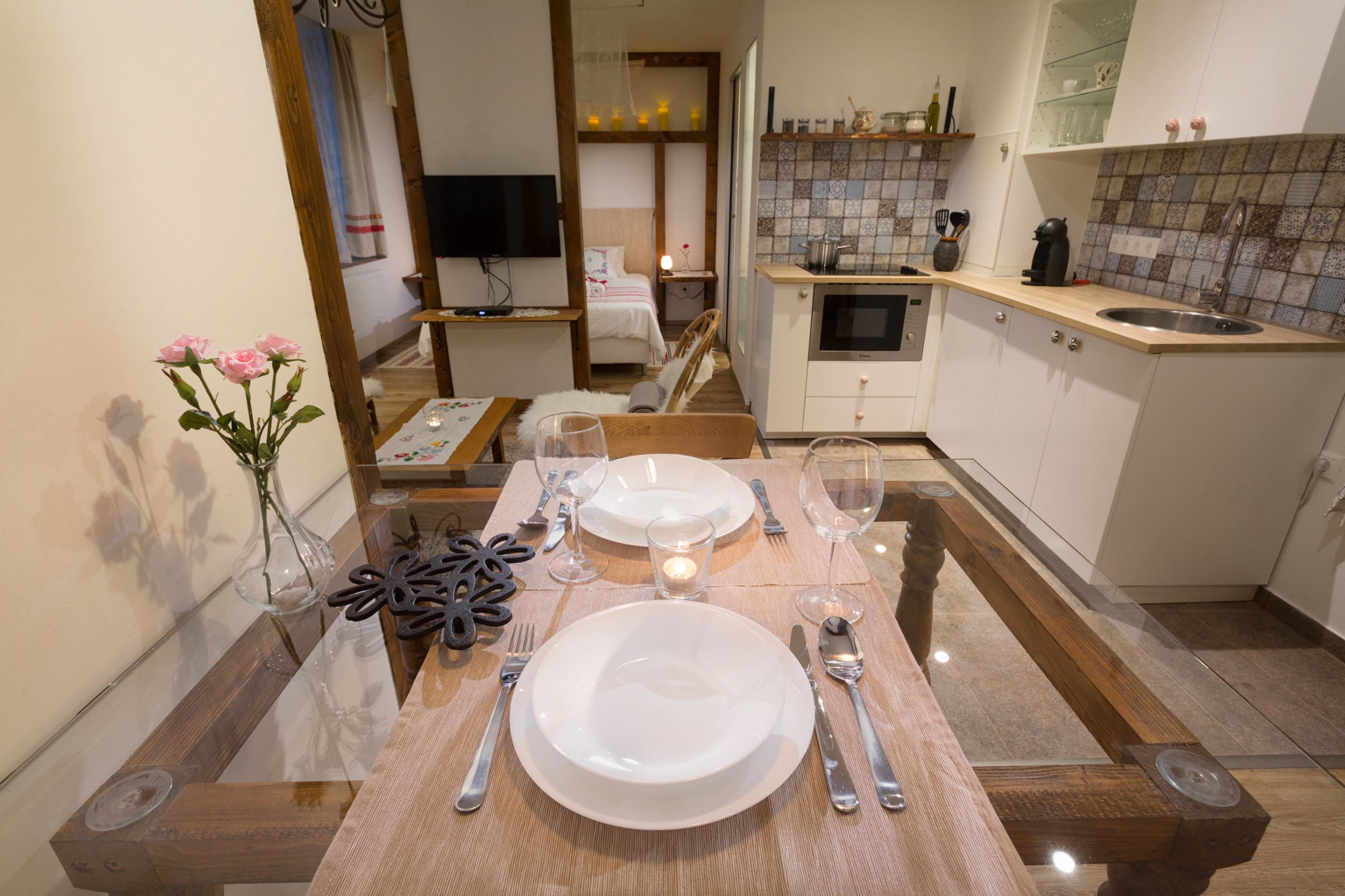 A dining area with a wooden table set for two, a vase with flowers, and a kitchen in the background.