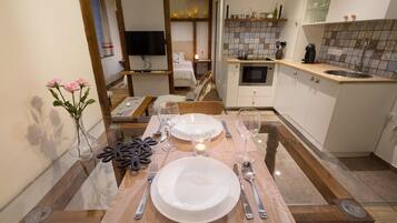 A dining area with a wooden table set for two, a vase with flowers, and a kitchen in the background.