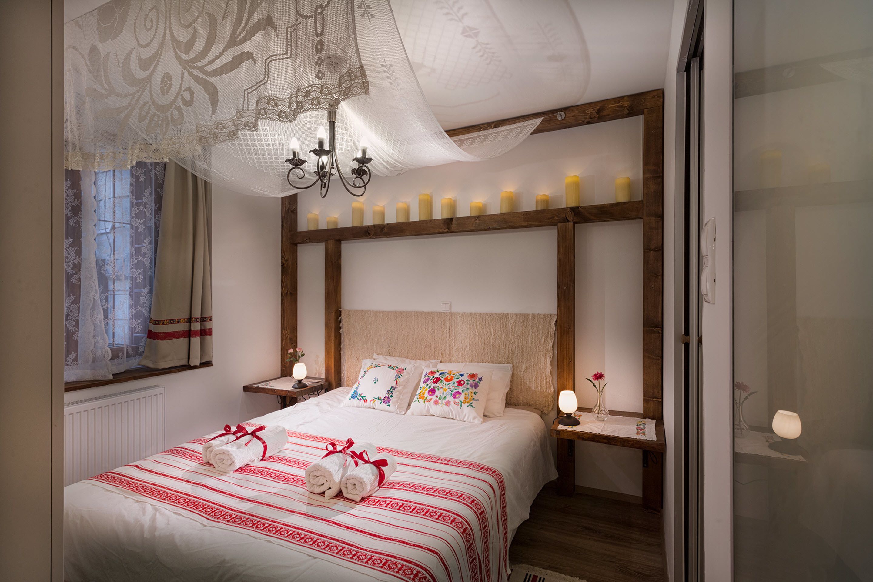 Een slaapkamer met een groot bed, decoratieve kussens, een kroonluchter, bedlampen en een raam met gordijnen.