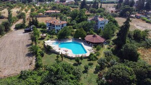 Eine Vogelperspektive zeigt eine Wohngegend mit einem großen Schwimmbecken, das von einem Garten, mehreren Häusern und einer hügeligen Landschaft umgeben ist.