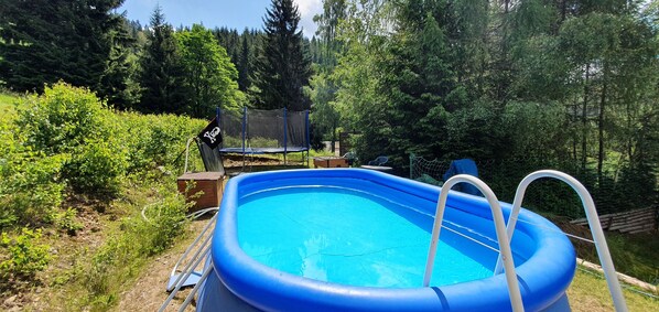 An outdoor swimming pool surrounded by a fence, with a trampoline and a chair nearby.