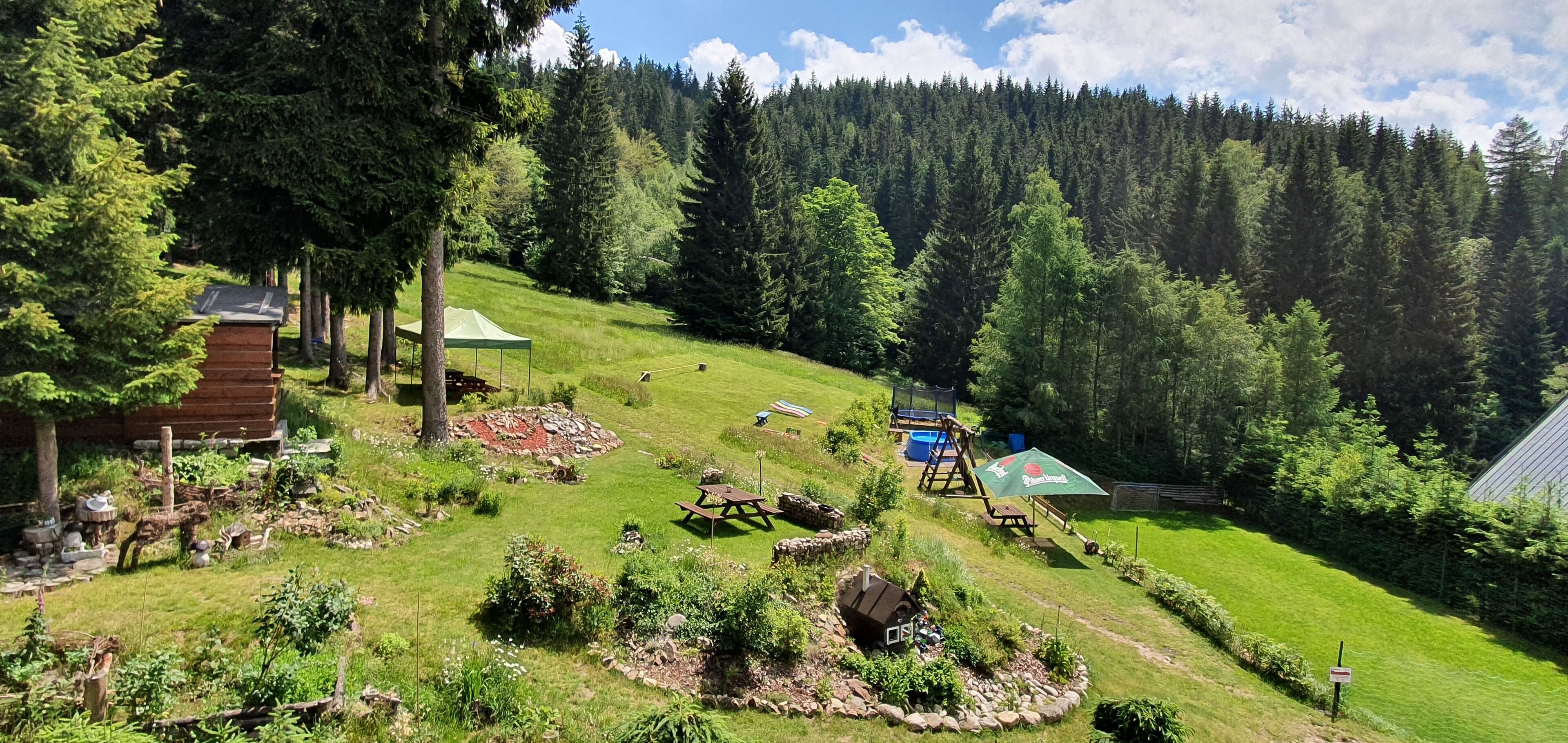 A rustic cabin with a green roof, surrounded by a lush garden and a forested hillside.