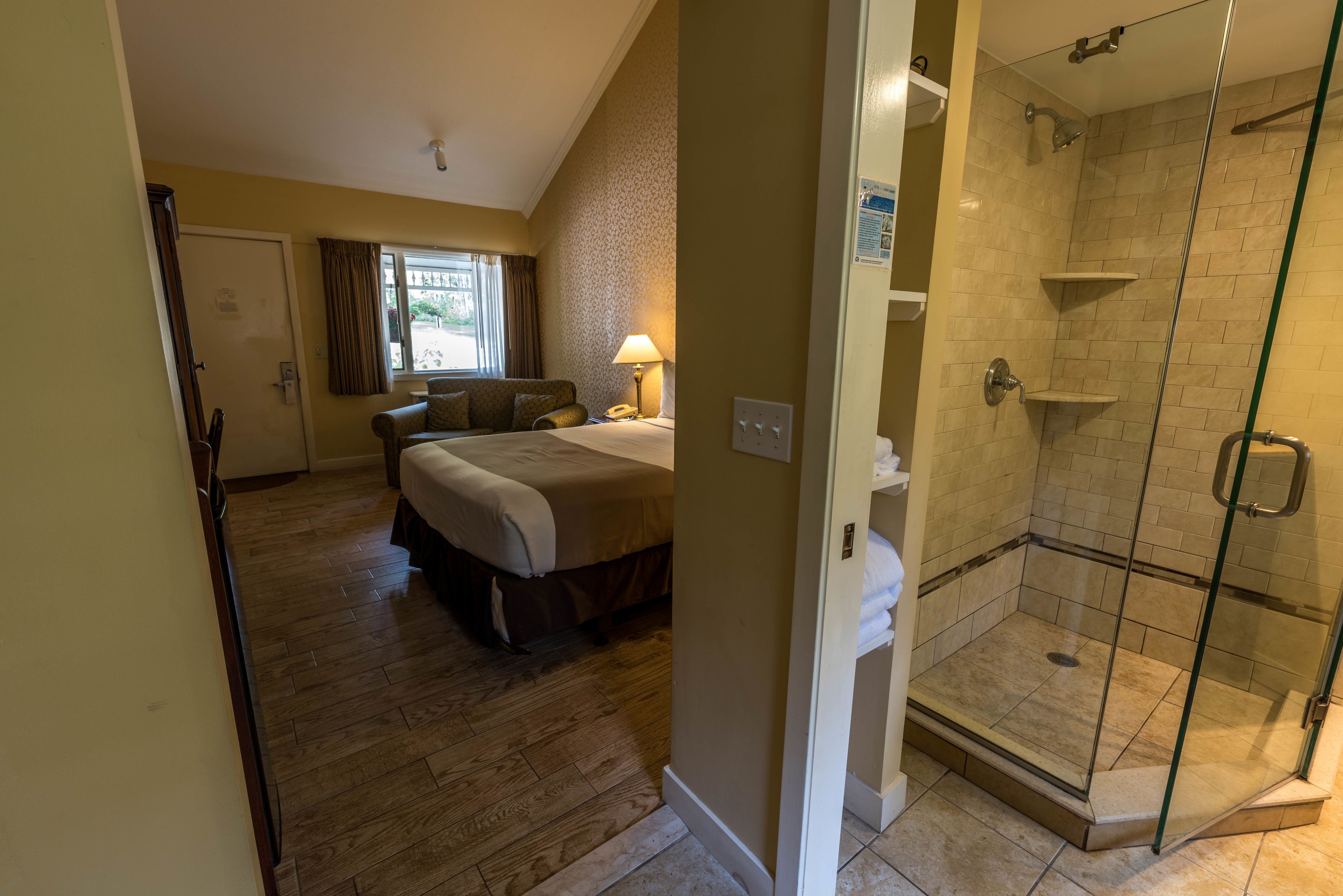A hotel room with a bed, a bedside table, a lamp, a shower area with glass doors, and a window with curtains.
