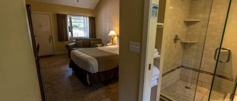 A hotel room with a bed, a bedside table, a lamp, a shower area with glass doors, and a window with curtains.