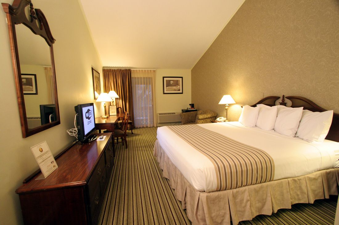Habitación de hotel con una cama grande, un escritorio con computadora, un espejo y una ventana con cortinas.