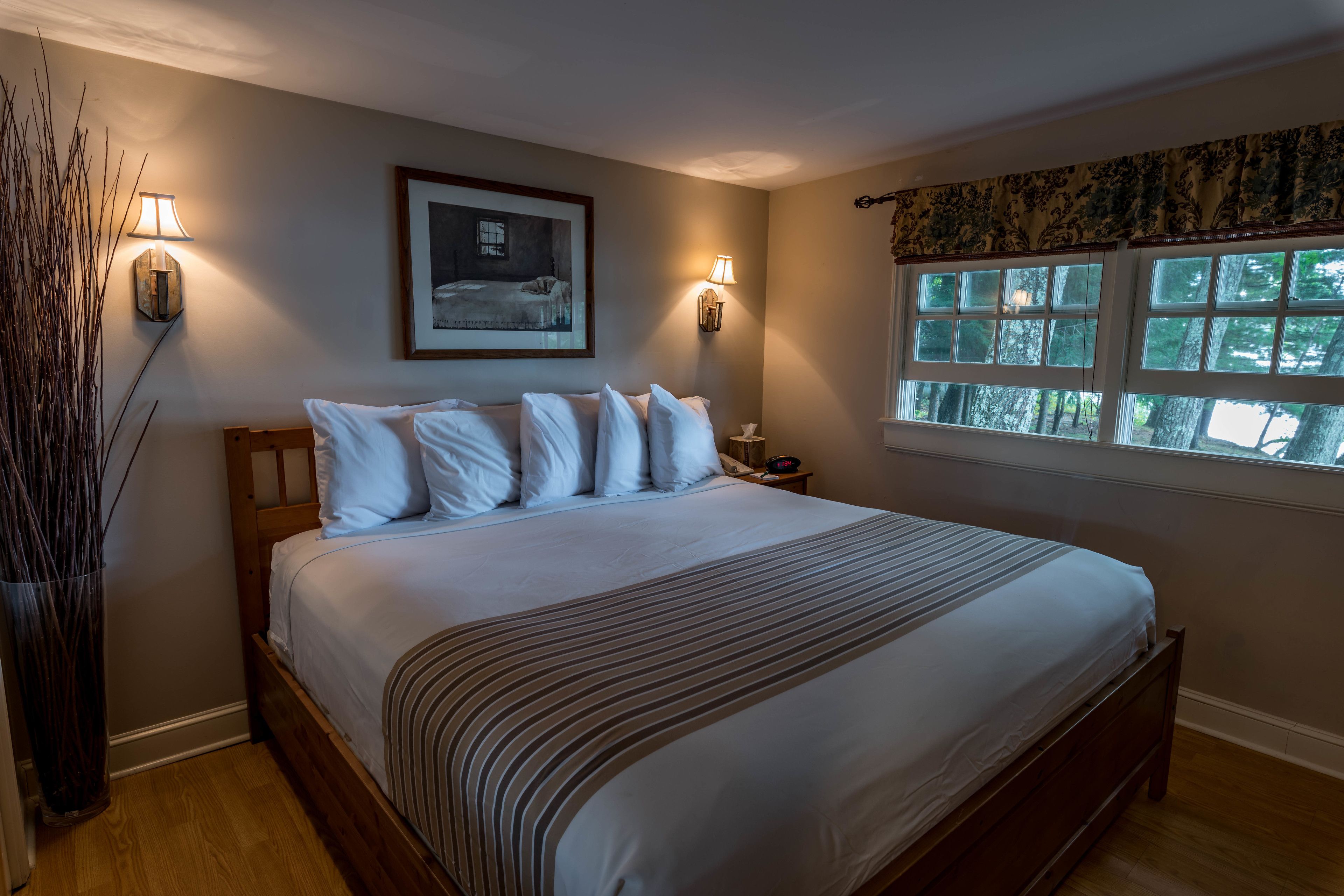 A bedroom with a large bed, two wall-mounted lamps, a framed picture, and a window with a view of trees.