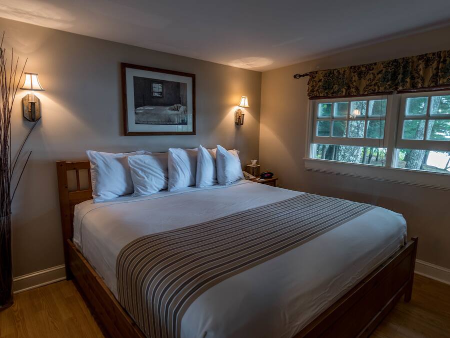 A bedroom with a large bed, two wall-mounted lamps, a framed picture, and a window with a view of trees.