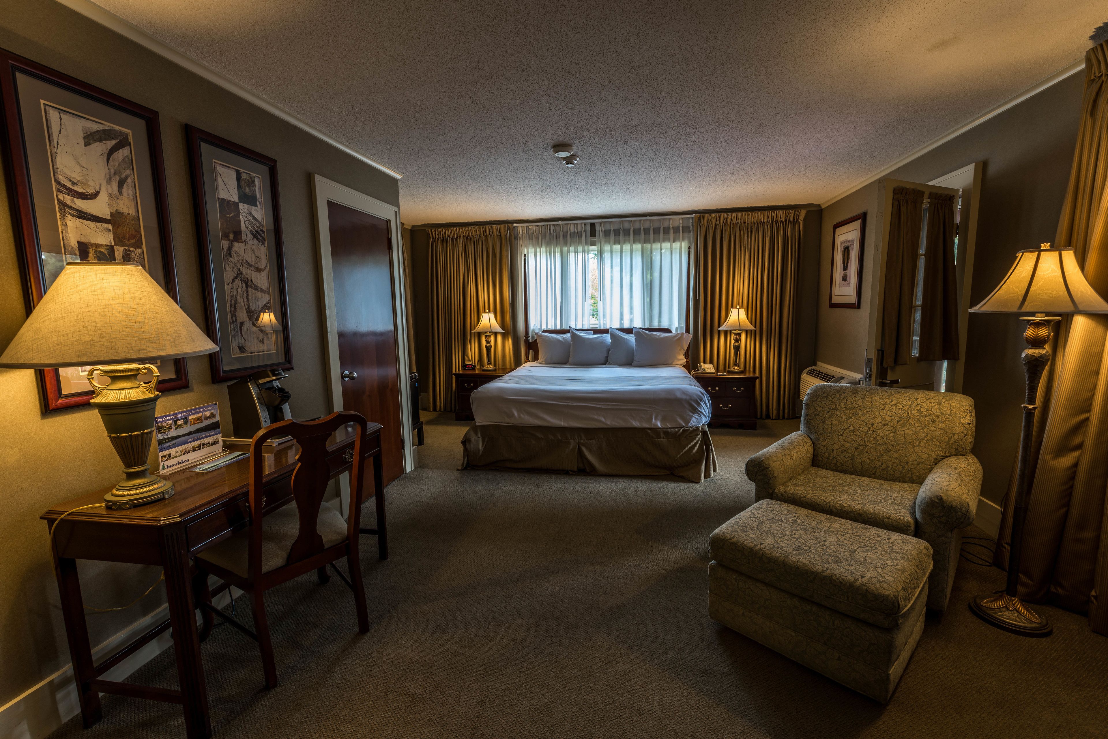 A hotel room with a bed, two bedside tables, a desk with a chair, a small armchair, and a floor lamp.