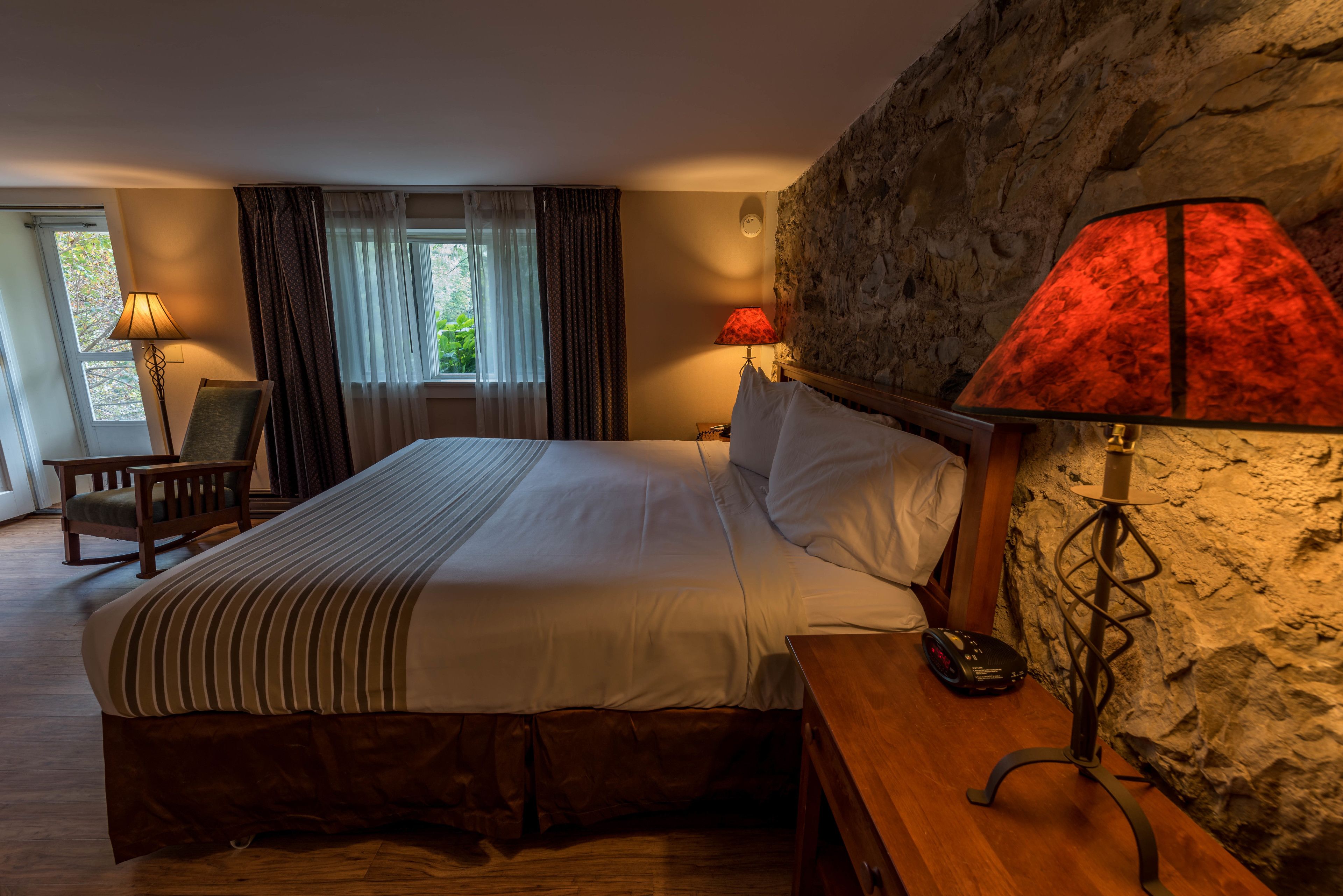 A hotel room with a large bed, two bedside lamps, a wooden nightstand, a chair, and a stone wall.