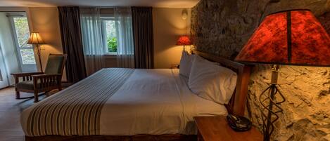 A hotel room with a large bed, two bedside lamps, a wooden nightstand, a chair, and a stone wall.