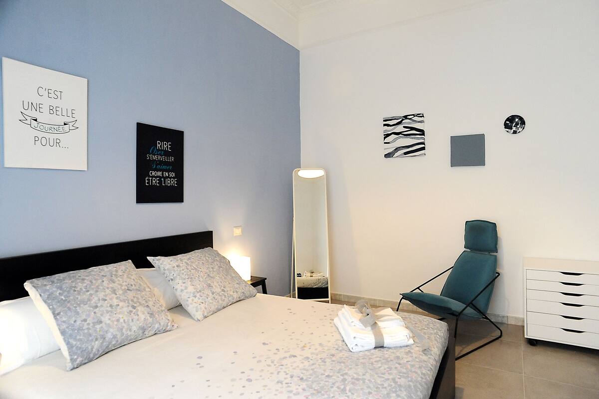 Ein ordentlich eingerichtetes Hotelzimmer mit Bett, Kissen, einem Sessel, einem Schminktisch und Wandbildern.