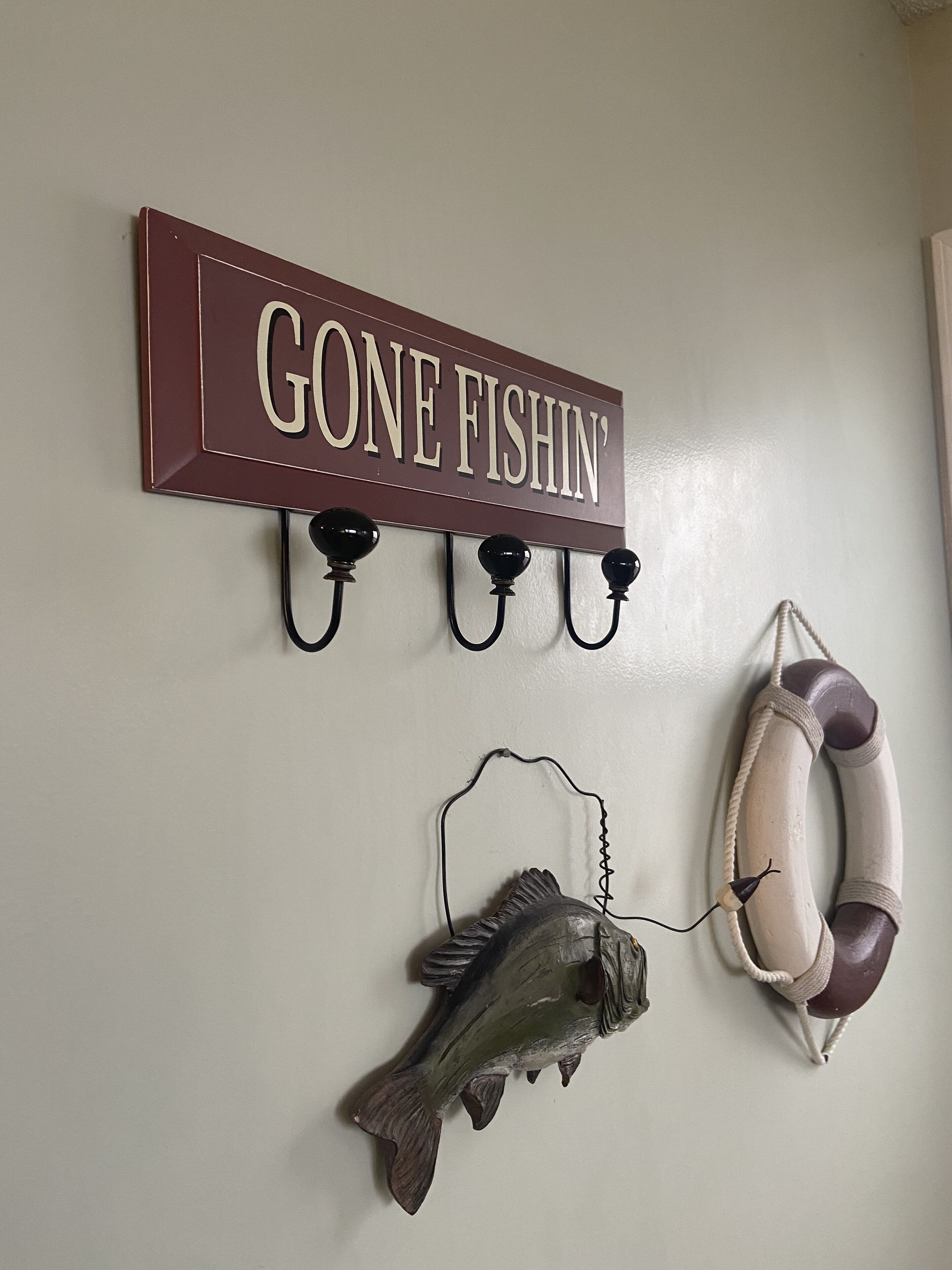 Un mur sur lequel est affiché un panneau indiquant « JE SUIS PARTI PÊCHER », ainsi que trois crochets ornés d’un motif en forme de poisson et d’un gilet de sauvetage.