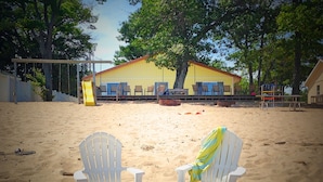 Dos sillas de playa blancas sobre una zona arenosa con un edificio al fondo.
