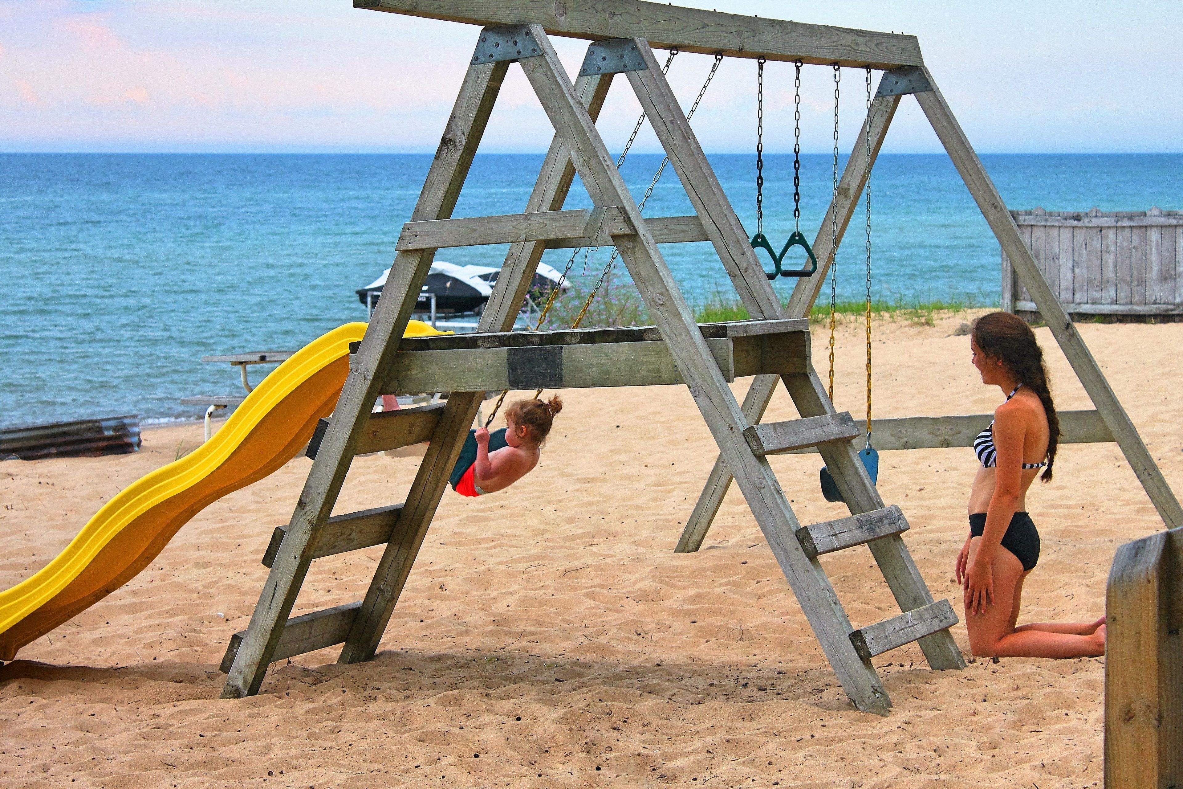 A wooden swing set with a yellow slide on a sandy beach with the ocean in the background.