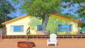 Una casa de playa con un árbol grande, una silla blanca y un barril metálico.