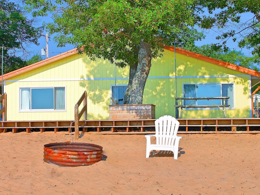 Une maison de plage avec un grand arbre, une chaise blanche et un baril en métal.