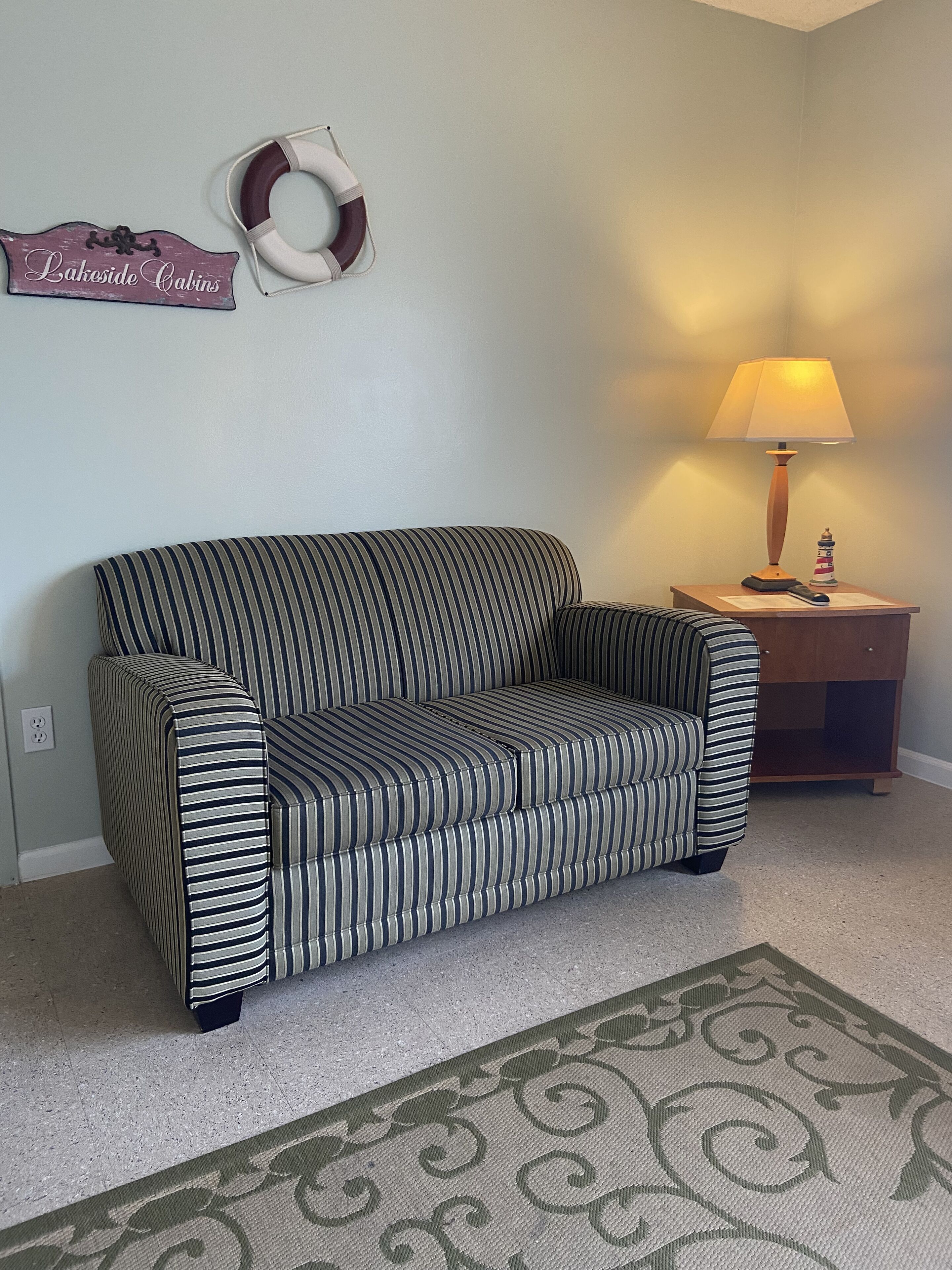 A striped sofa in a room with a wall-mounted sign reading "Lakeside Cabins," a small table with a lamp, and a decorative rug.