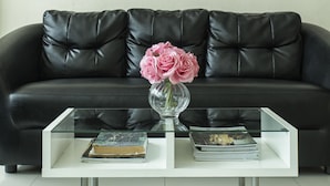 A living room with a black leather sofa, a glass coffee table with books and a vase of flowers, and a flat-screen TV mounted on the wall. - Kulasab Hotel (Hat Yai)