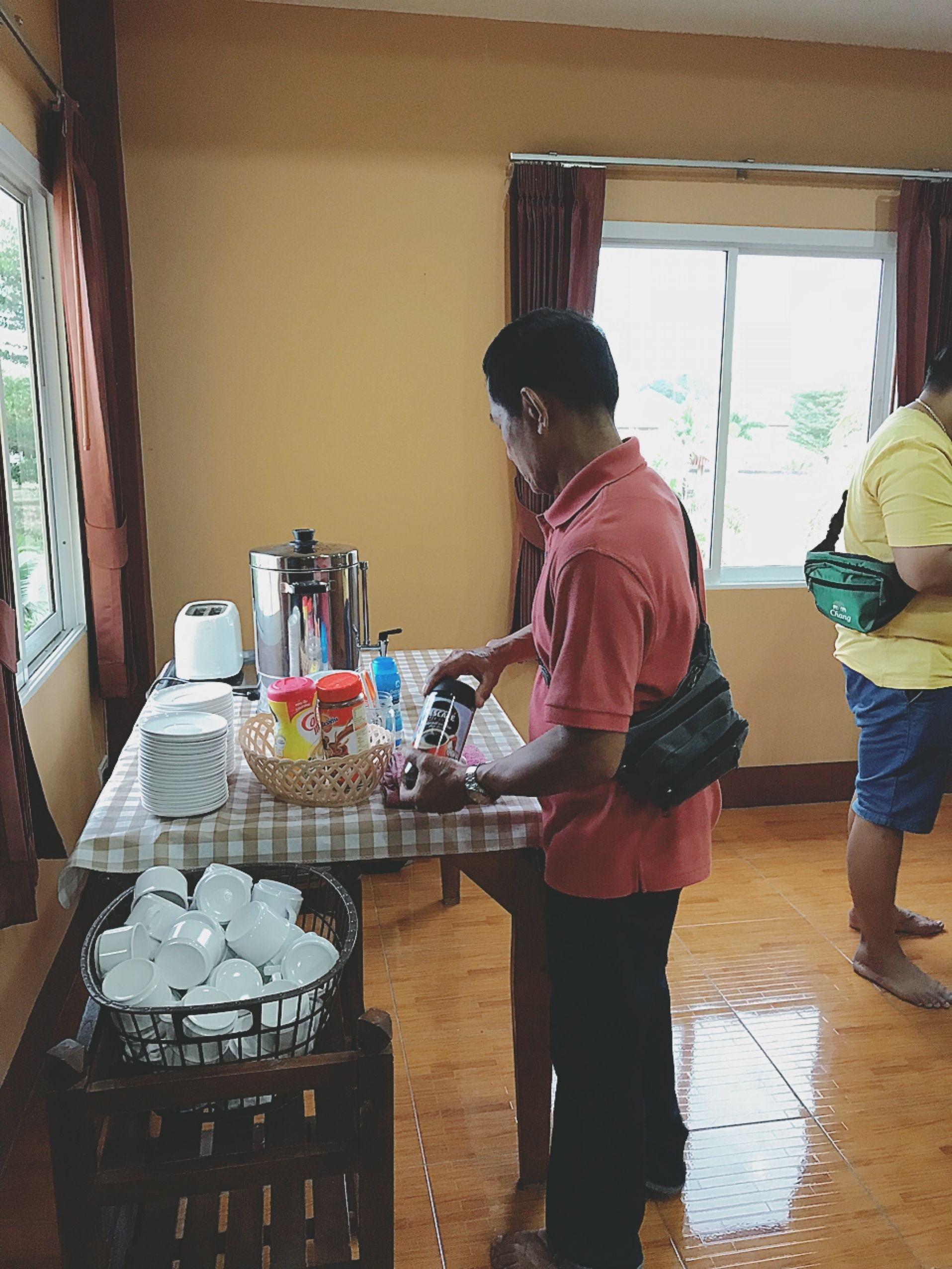 Una habitación con una mesa dispuesta para una comida, una ventana con cortinas y una persona preparando alimentos.