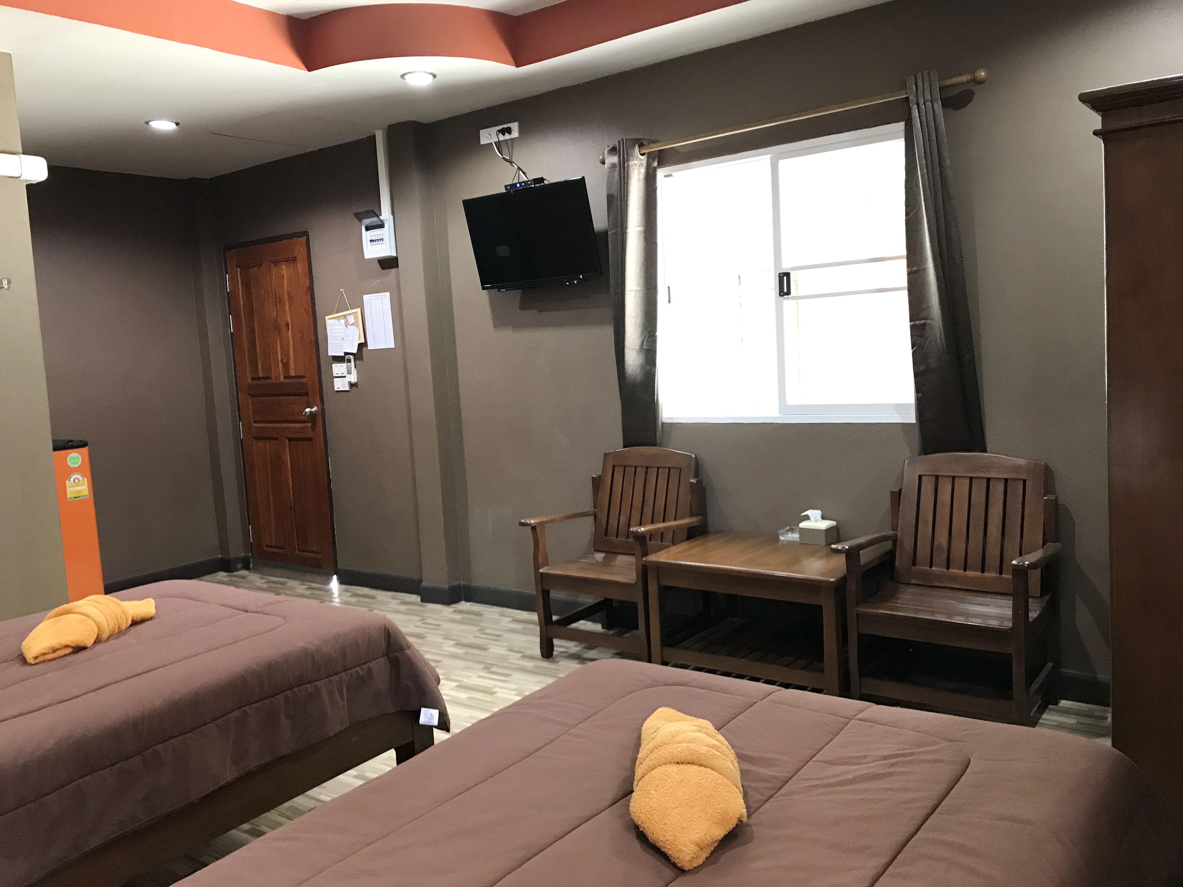 Habitación de hotel con dos camas, una silla de madera, una mesita, un televisor montado en la pared y un ventanal con cortinas.