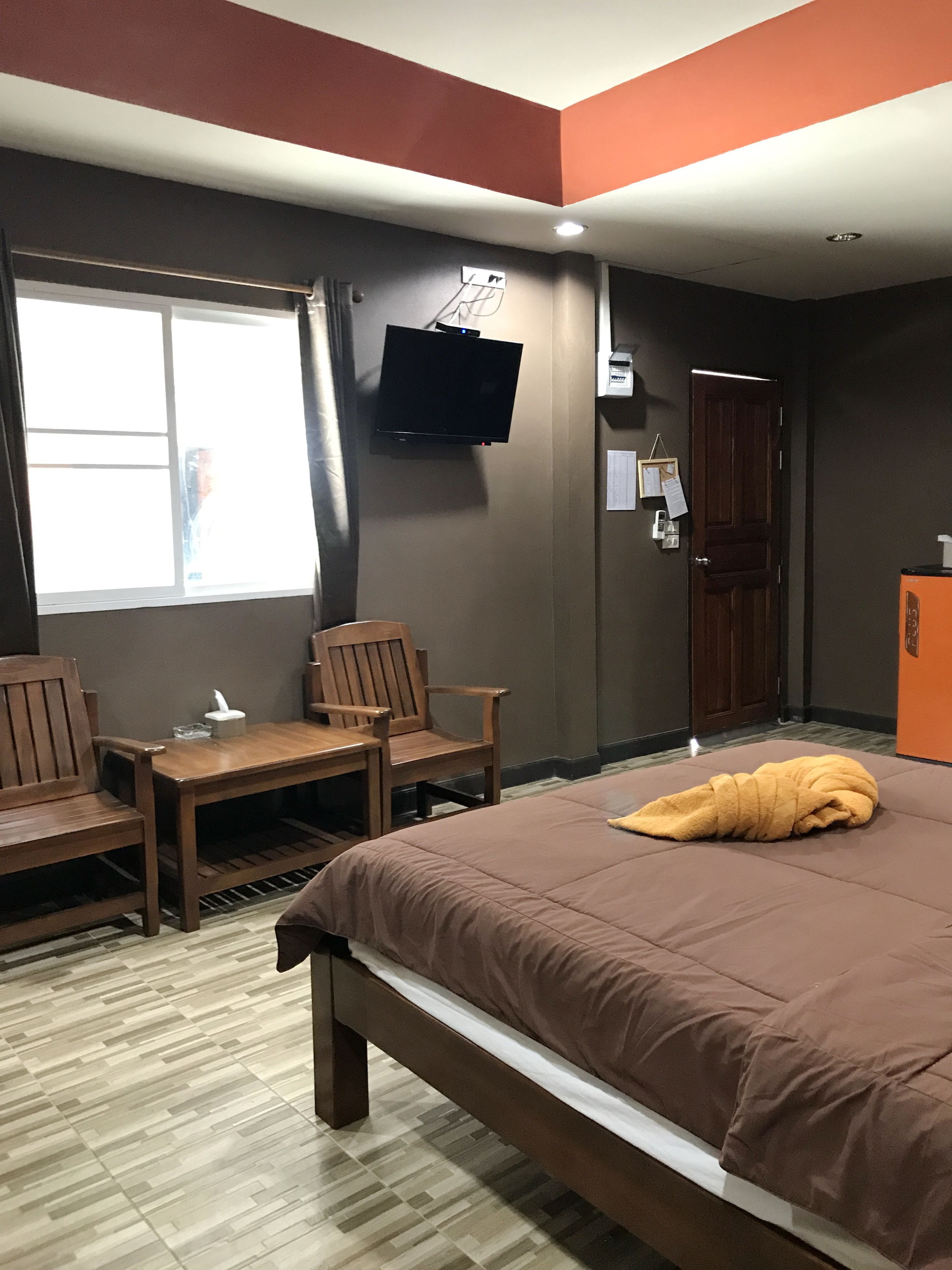 A hotel room with a bed, two wooden chairs, a small table, a TV mounted on the wall, and a window with curtains.