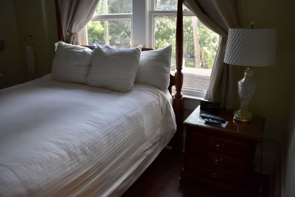 A neatly made bed with white linens and pillows, a bedside table with a lamp and remote control, and a window with curtains.