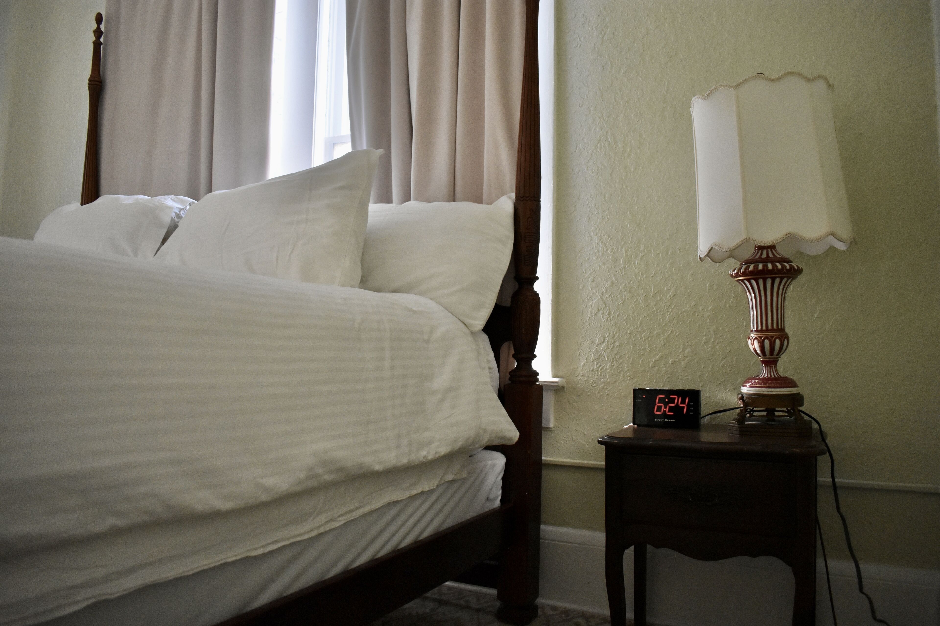 A neatly made bed with white linens, a wooden headboard, and a bedside table with a lamp and a digital clock displaying the time.