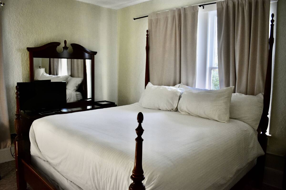 A bedroom with a large bed, a wooden headboard, a nightstand, a mirror, and a window with curtains.