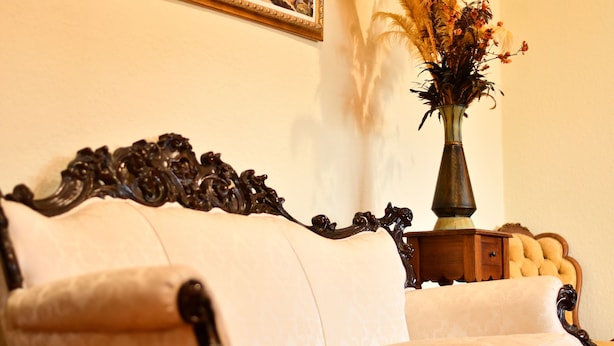 A room with a white upholstered sofa, a dark wooden side table with a vase of dried plants, and a framed painting on the wall. - The DeLand Hotel (DeLand)