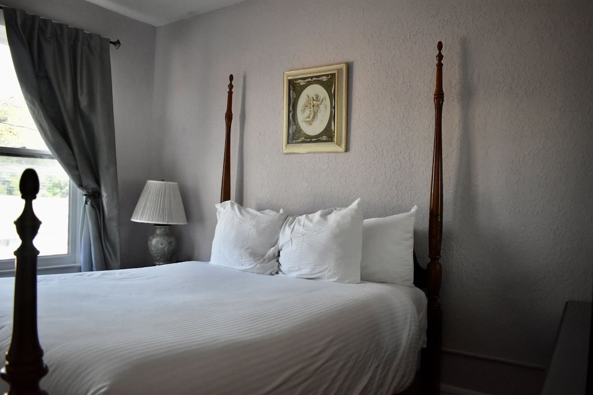A neatly made bed with white linens and two pillows, a bedside lamp, and a framed artwork on the wall.