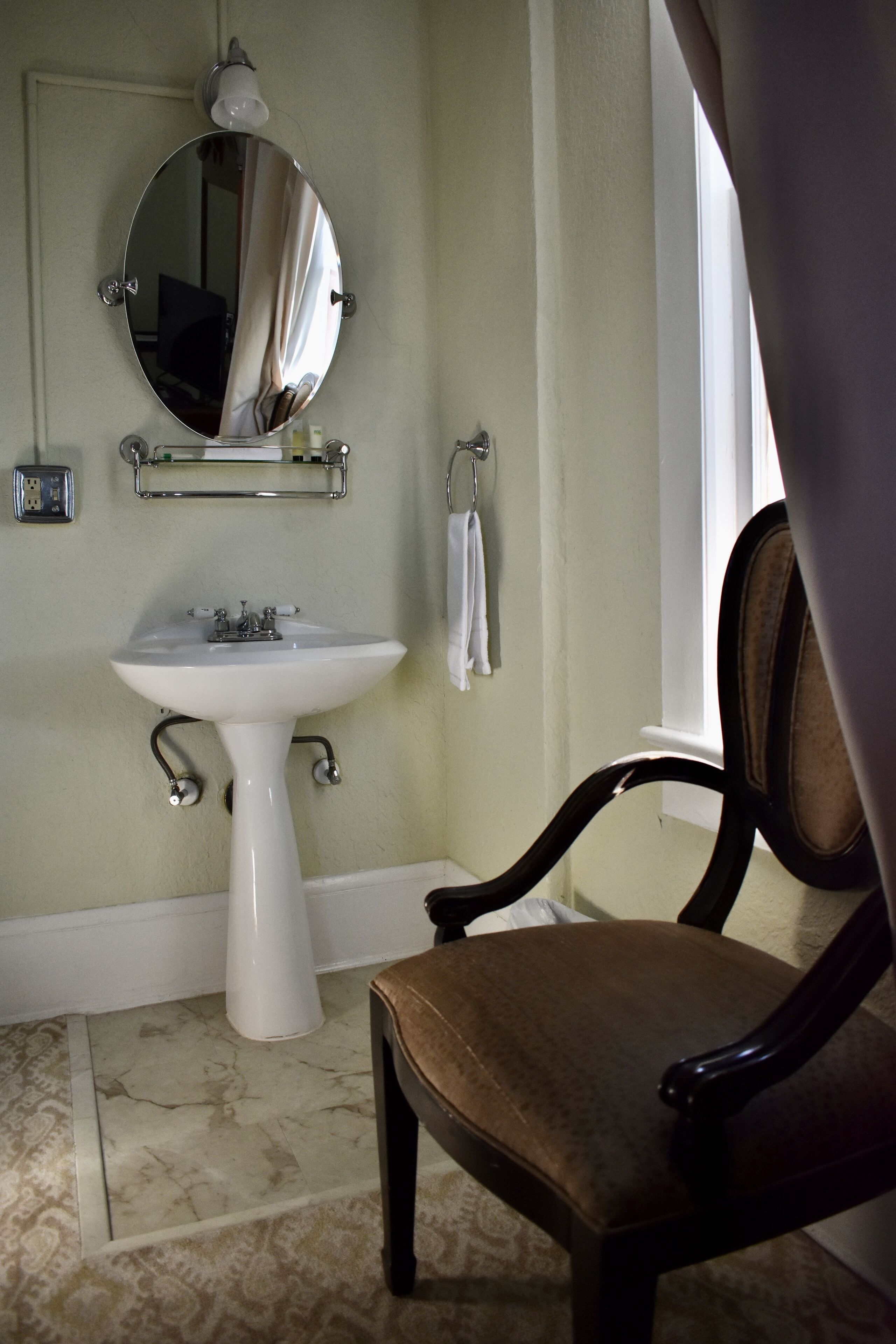 A bathroom with a pedestal sink, a mirror, a towel rack, and a chair.