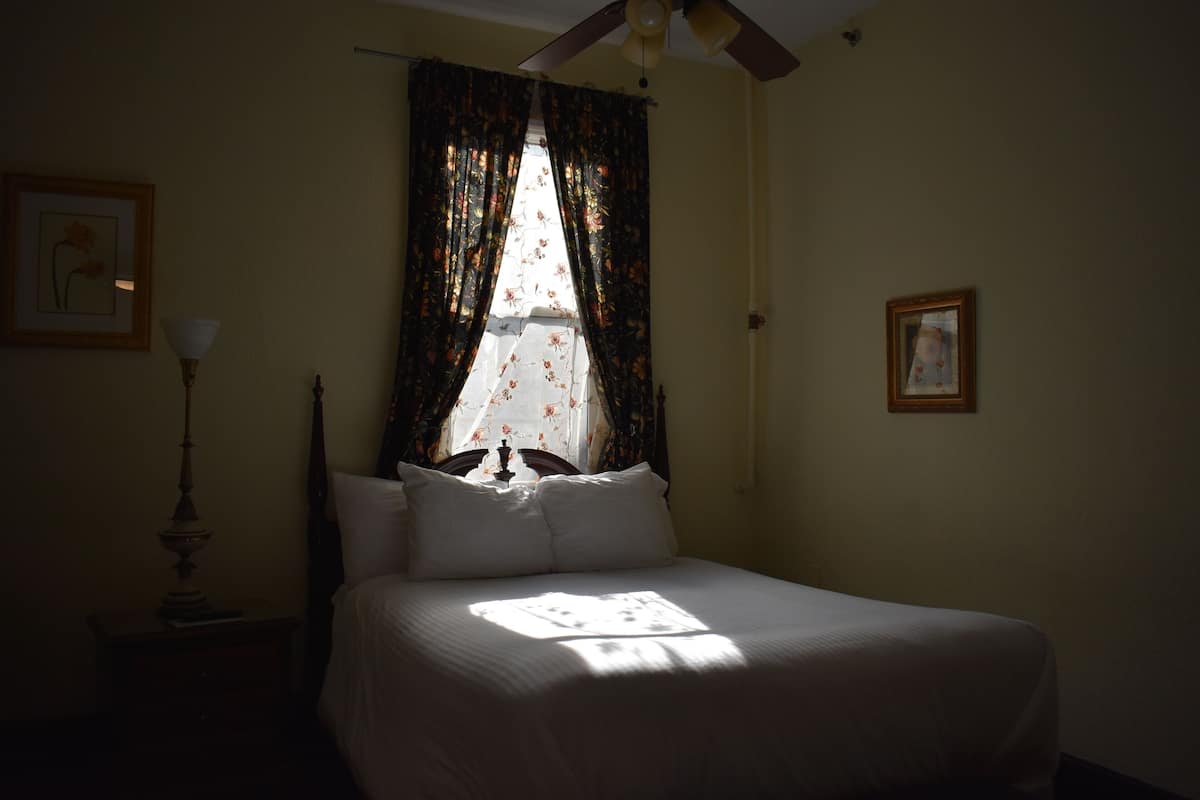 A bedroom with a bed, a ceiling fan, a window with floral curtains, and a nightstand with a lamp.