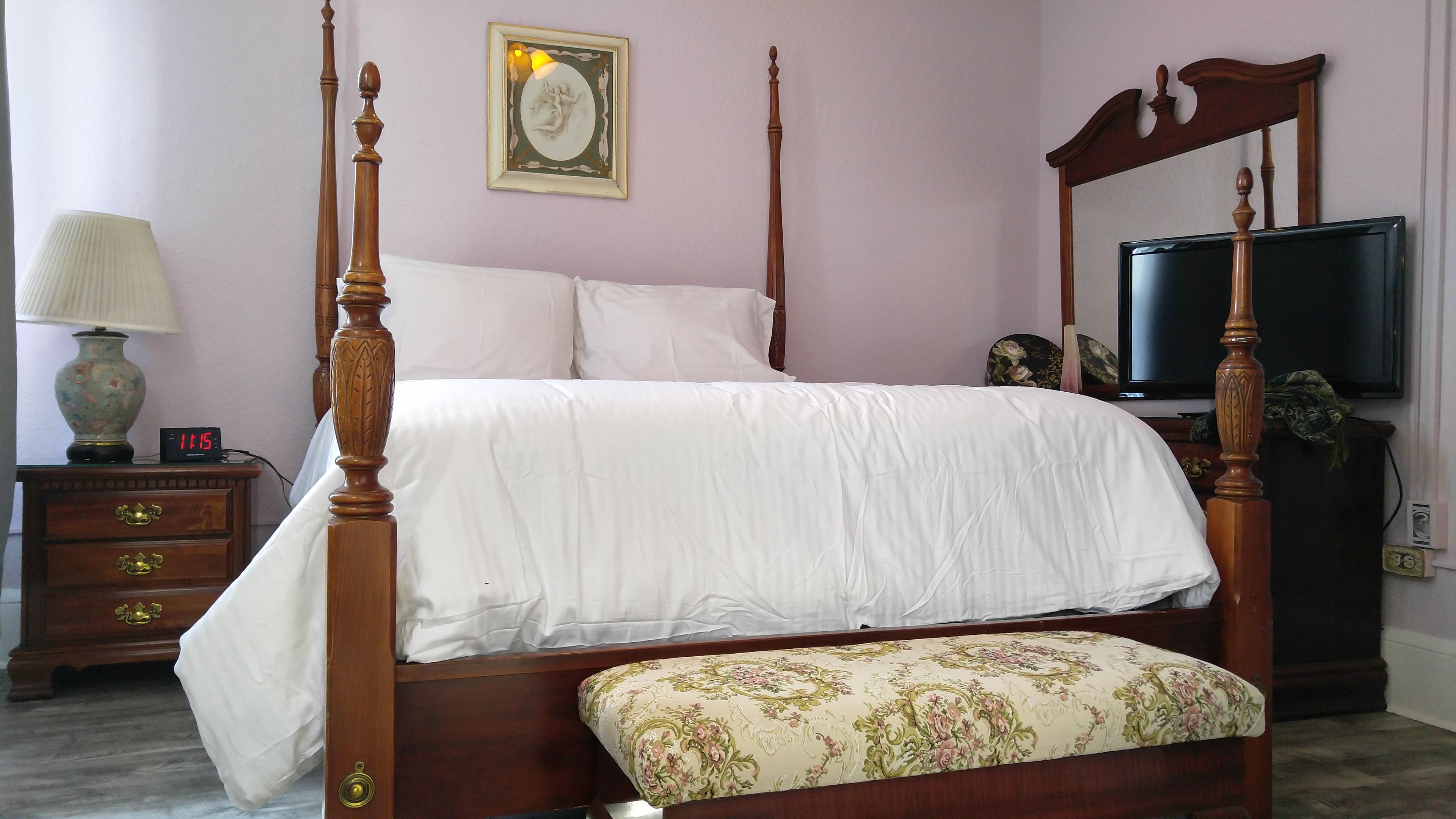 Una cama con dosel, un banco floral, una mesita de noche con lámpara y un televisor sobre un mueble.