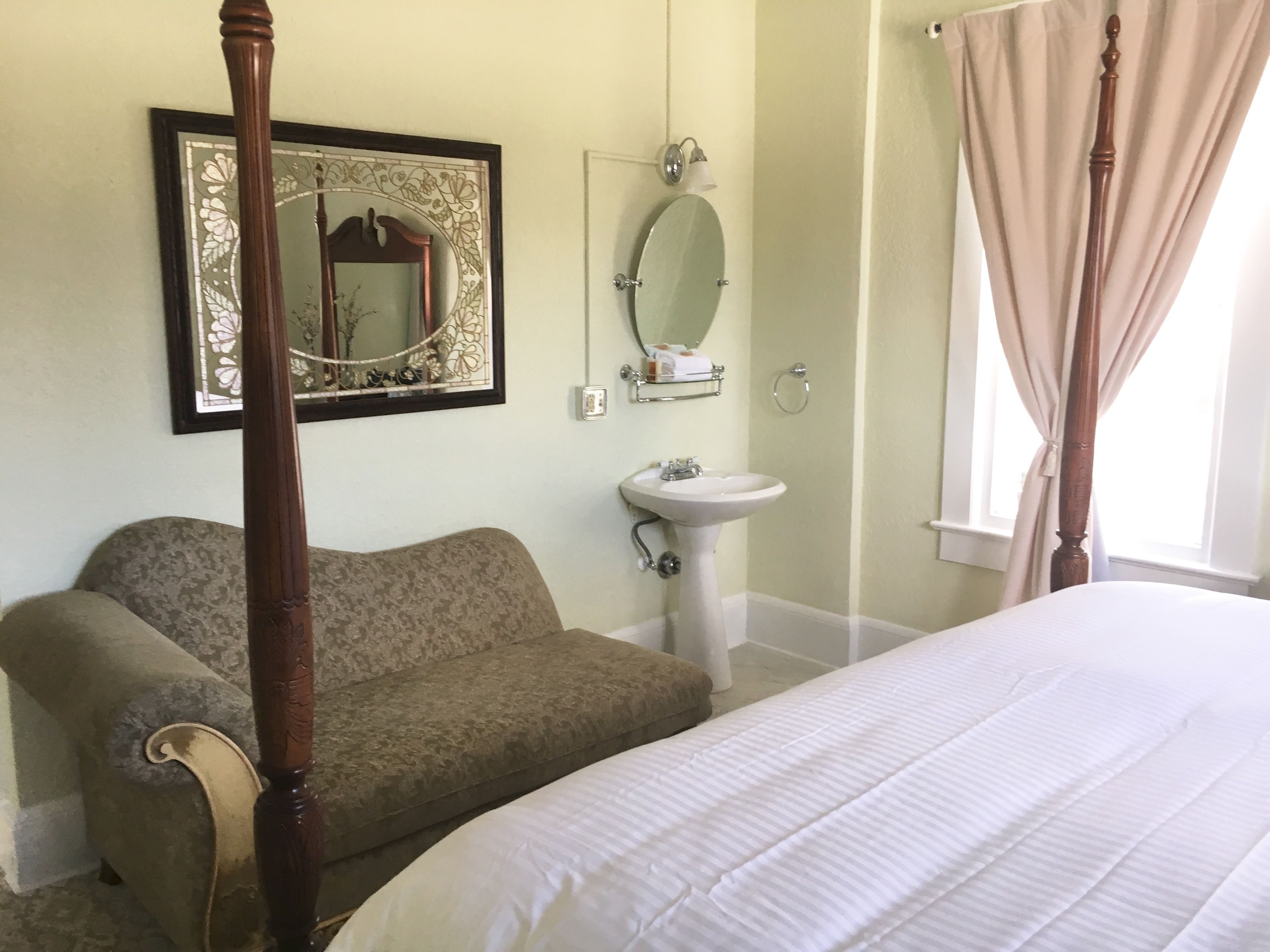 Quarto com cama de dossel, sofá, mesa de cabeceira, espelho e janela com cortinas.