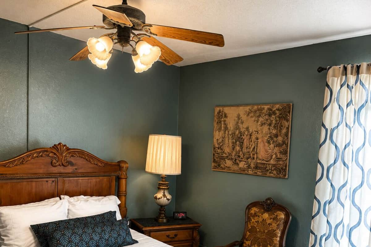A bedroom with a wooden bed, a nightstand with a lamp, a chair, a ceiling fan, and a painting on the wall.