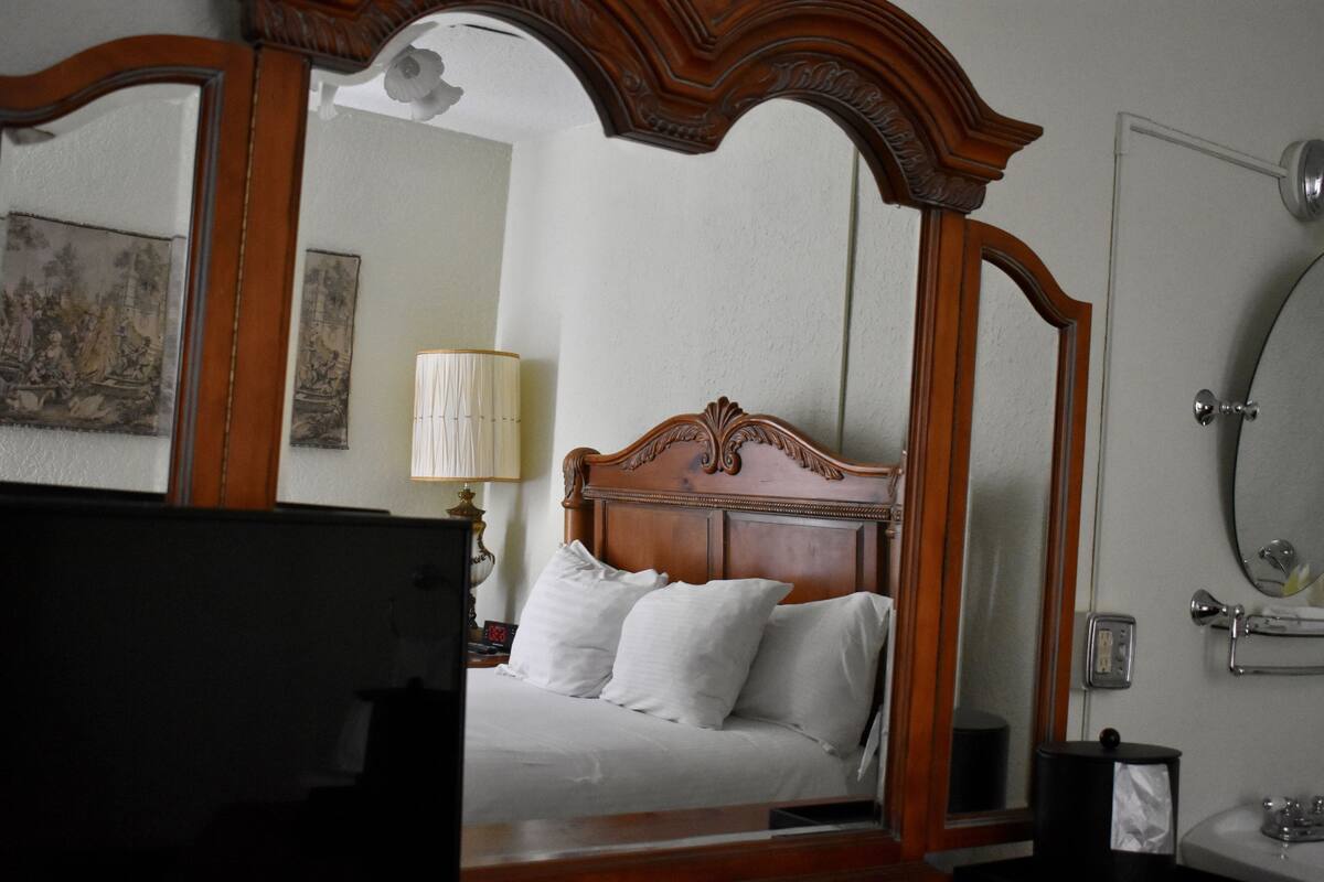 A bedroom with a wooden four-poster bed, a bedside table with a lamp, a mirror, and a small nightstand.