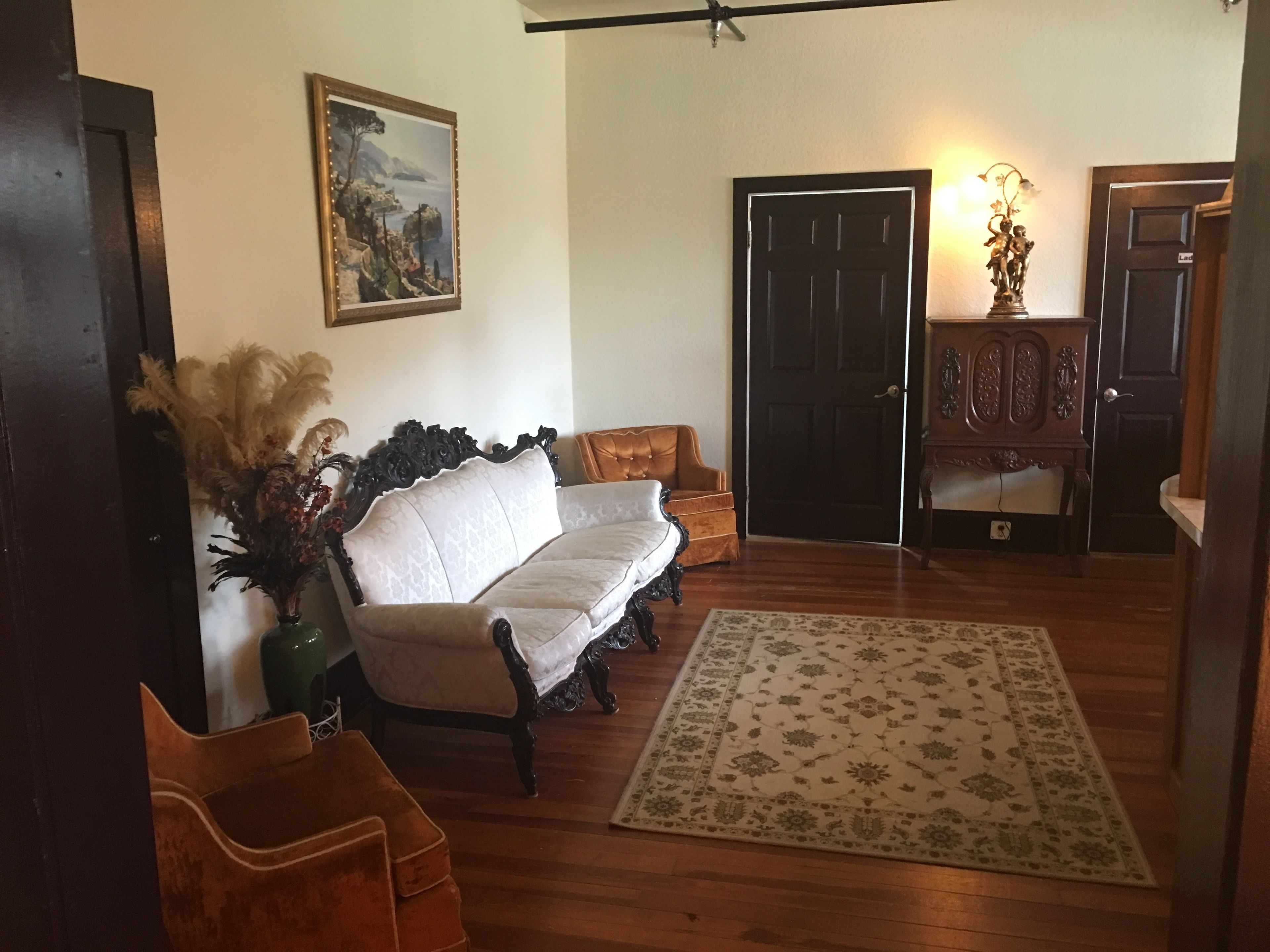 Un salon avec un canapé blanc, un plancher en bois, un tapis à motifs, un tableau accroché au mur et une porte dotée d’un luminaire.